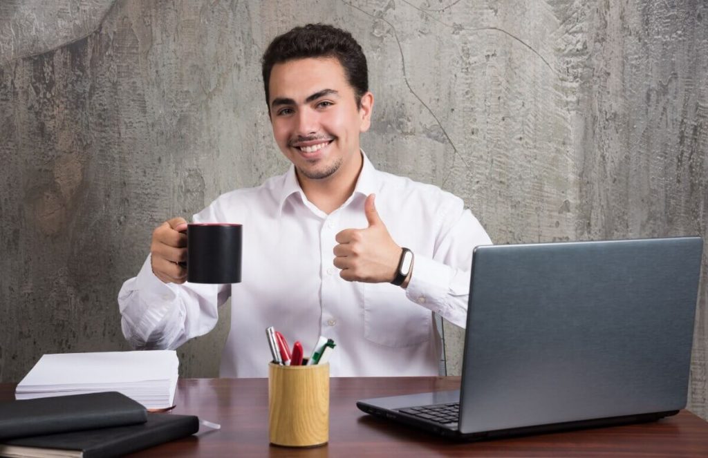 Businessman-holding-cup-tea-showing-thumbs-up-office-desk